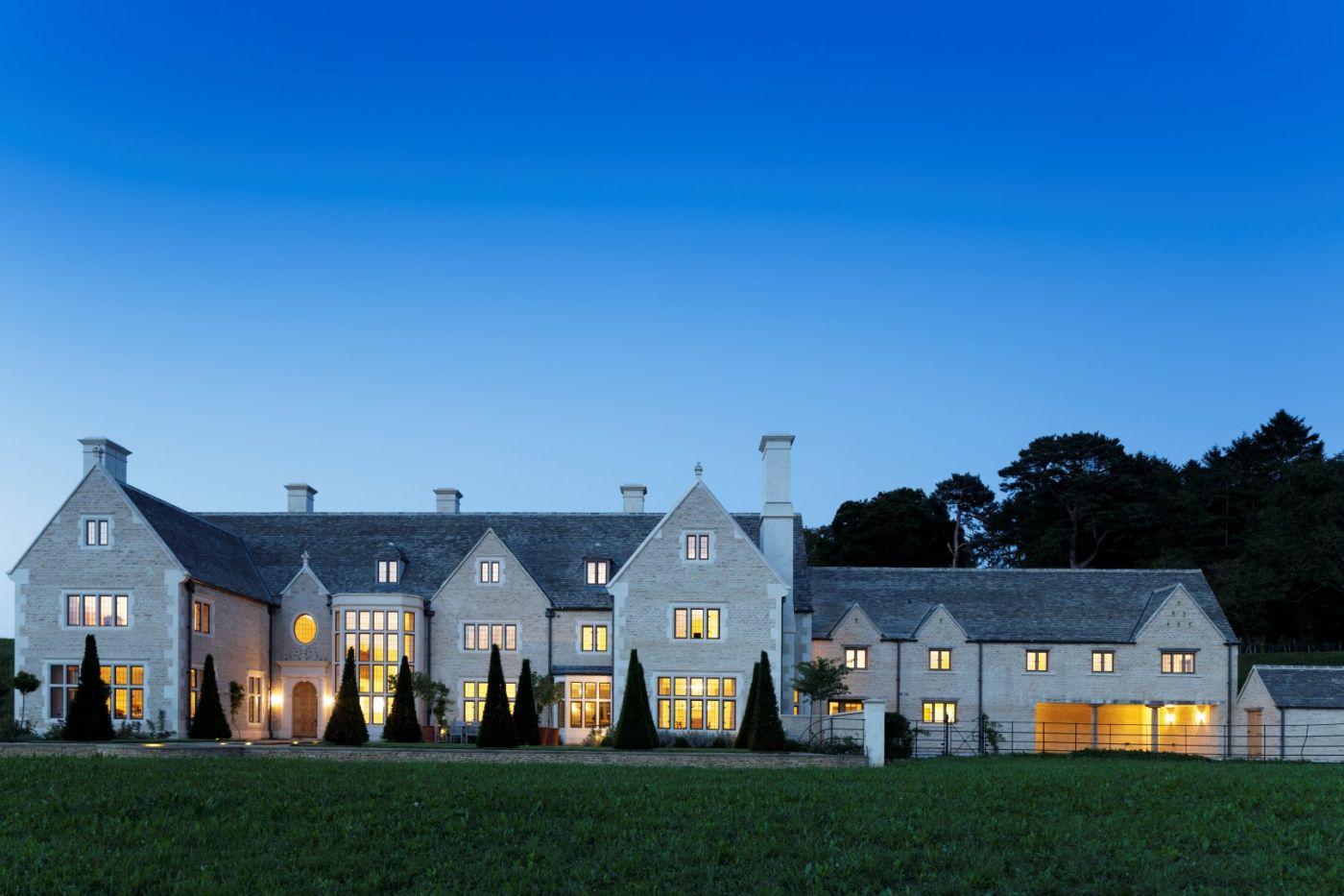 A wide shot of a large, modern Cotswold home in Church Enstone at dusk. There are lights on in every room of the home.